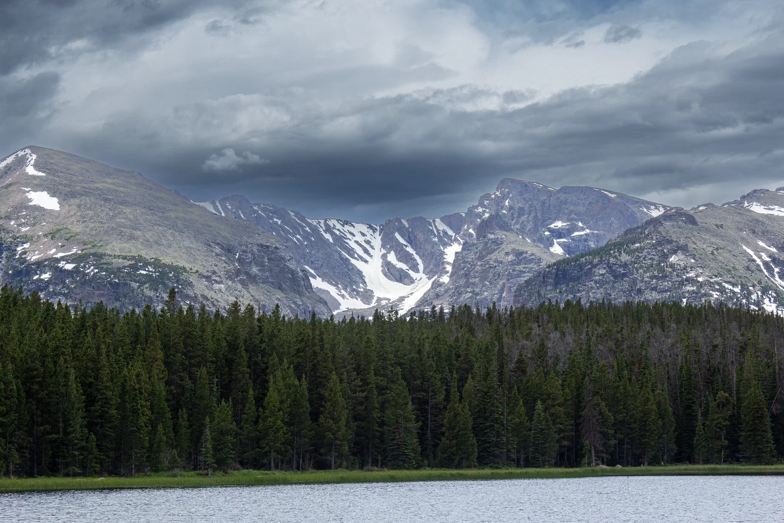 Snow-streaked peaks above a dense pine forest and a lake under storm clouds